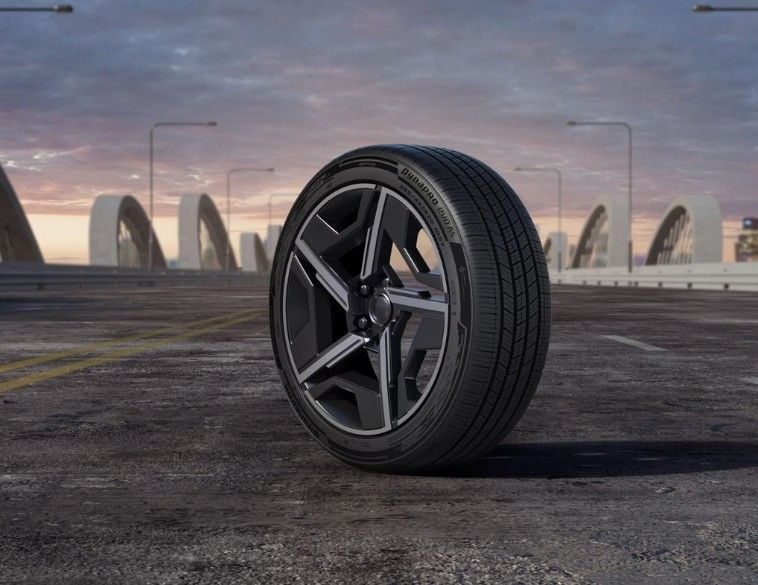 Un pneu Hankook Dynapro Evo AS monté sur une jante moderne noire et argentée, positionné sur une route asphaltée avec un pont et un ciel au crépuscule en arrière-plan.