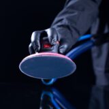 A close-up of a technician's gloved hand using a 3M power sander equipped with a blue net abrasive disc and a dust extraction vacuum hose.