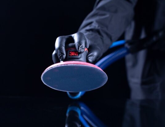 A close-up of a technician's gloved hand using a 3M power sander equipped with a blue net abrasive disc and a dust extraction vacuum hose.