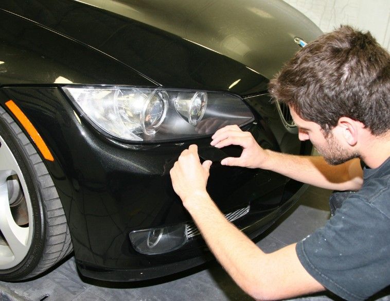 A technician in a black t-shirt performs a cosmetic repair on the front bumper of a black BMW.