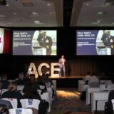 A keynote speaker stands on stage at the Automotive Conference & Expo (ACE) in front of an audience of automotive professionals. The stage features large, shimmering silver letters spelling "ACE" and is flanked by two large screens displaying a presentation by Paul Smith, Commander of the Royal Canadian Navy, titled "Navigating Change: Command Leadership Beyond the Playbook." A Canadian flag hangs to the left of the stage. In the foreground, attendees are seated at tables with chair covers.