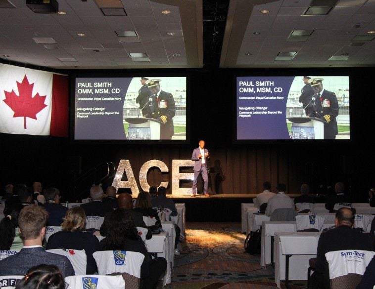 A keynote speaker stands on stage at the Automotive Conference & Expo (ACE) in front of an audience of automotive professionals. The stage features large, shimmering silver letters spelling "ACE" and is flanked by two large screens displaying a presentation by Paul Smith, Commander of the Royal Canadian Navy, titled "Navigating Change: Command Leadership Beyond the Playbook." A Canadian flag hangs to the left of the stage. In the foreground, attendees are seated at tables with chair covers.