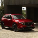 Un tout nouveau Mazda CX-5 2026 rouge mettant en valeur l'évolution du design Kodo, stationné dans une entrée en gravier devant un bâtiment moderne en bois foncé. Le VUS est doté de roues noires et d'une silhouette profilée et aérodynamique.