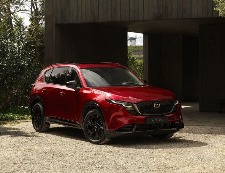 Un tout nouveau Mazda CX-5 2026 rouge mettant en valeur l'évolution du design Kodo, stationné dans une entrée en gravier devant un bâtiment moderne en bois foncé. Le VUS est doté de roues noires et d'une silhouette profilée et aérodynamique.