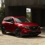 A red all-new 2026 Mazda CX-5 showcasing the evolved Kodo Design parked on a gravel driveway in front of a modern dark wood building. The SUV features black wheels and a sleek, aerodynamic silhouette.
