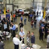 An overhead view of a busy auto parts trade show in a warehouse, featuring manufacturer booths, brand signage like Bestbuy, and people networking and learning.