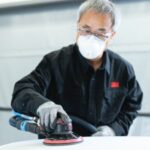 Technicien en carrosserie utilisant une ponceuse avec système d'aspiration, portant un équipement de protection individuelle complet (masque respiratoire, lunettes et gants).
