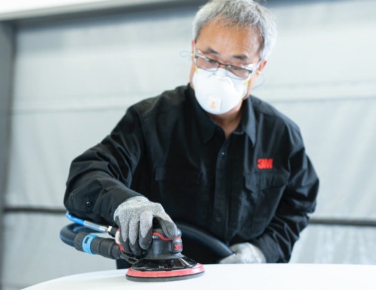 Technicien en carrosserie utilisant une ponceuse avec système d'aspiration, portant un équipement de protection individuelle complet (masque respiratoire, lunettes et gants).