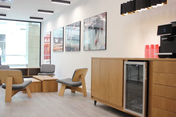 L’aire de détente moderne à l’intérieur du carrefour client Clutch au centre commercial Bayshore, comprenant du mobilier en bois clair, des fauteuils rembourrés gris, un coin café avec des gobelets rouges à l’effigie de Clutch et de grandes photos d’automobiles encadrées sur un mur blanc.