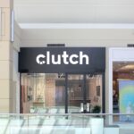 A bright storefront for the Clutch Customer Hub located inside the Bayshore Shopping Centre in Ottawa. The shop features a minimalist design with a prominent black sign displaying the white "clutch" logo above glass doors. The interior shows a clean, modern space with computers for guided digital car shopping.