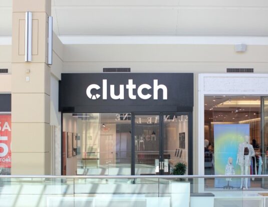 A bright storefront for the Clutch Customer Hub located inside the Bayshore Shopping Centre in Ottawa. The shop features a minimalist design with a prominent black sign displaying the white "clutch" logo above glass doors. The interior shows a clean, modern space with computers for guided digital car shopping.