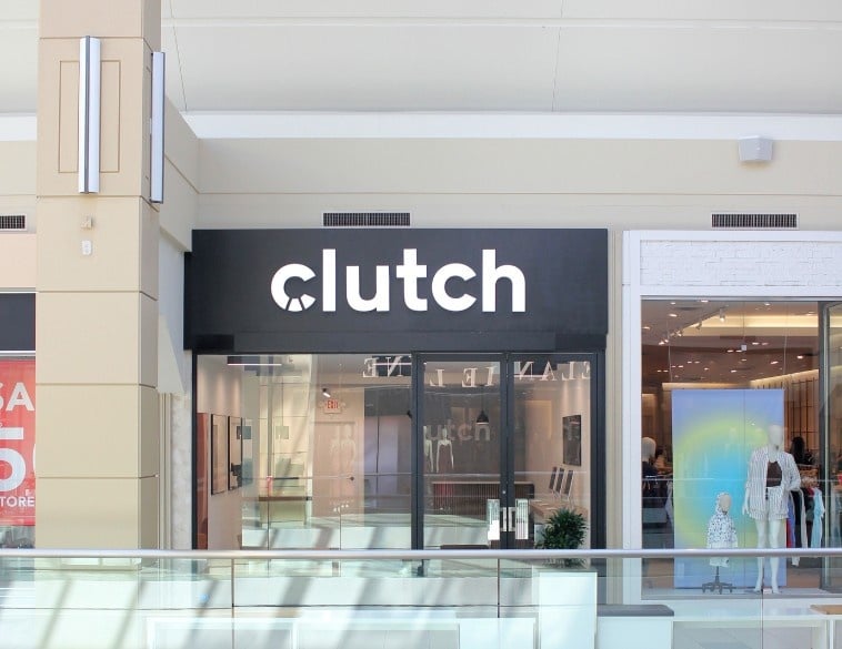 A bright storefront for the Clutch Customer Hub located inside the Bayshore Shopping Centre in Ottawa. The shop features a minimalist design with a prominent black sign displaying the white "clutch" logo above glass doors. The interior shows a clean, modern space with computers for guided digital car shopping.