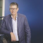 Un portrait de Jean Novak, président d'Unimax, portant des lunettes et une veste écossaise, appuyé contre une rampe d'escalier sur un fond bleu.