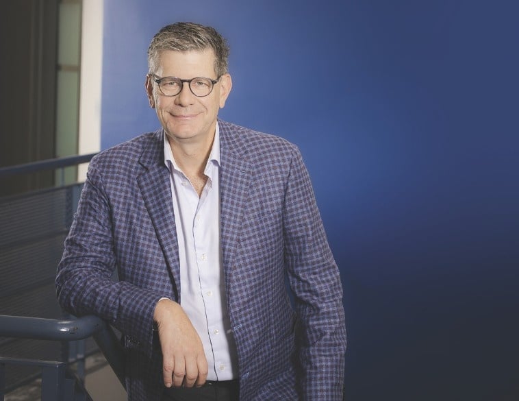Un portrait de Jean Novak, président d'Unimax, portant des lunettes et une veste écossaise, appuyé contre une rampe d'escalier sur un fond bleu.