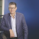 A portrait of Jean Novak, President and Chief Executive Officer of Unimax , wearing glasses and a checkered blazer while leaning against a railing in front of a blue background.