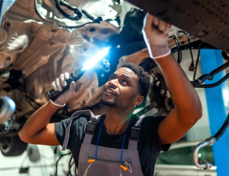 An automotive technician using a handheld flashlight to inspect the undercarriage of a vehicle in a service center.