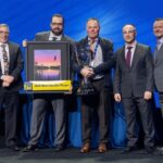 Brian Adcock, propriétaire de Fountain Tire d’Innisfail, se tient sur scène avec quatre autres hommes, tenant un grand trophée en argent. À ses côtés, un dirigeant tient un prix encadré de « Joueur le plus utile 2025 » arborant un logo « 70 ans. » Ce prix a été remis lors du congrès des propriétaires de Fountain Tire.