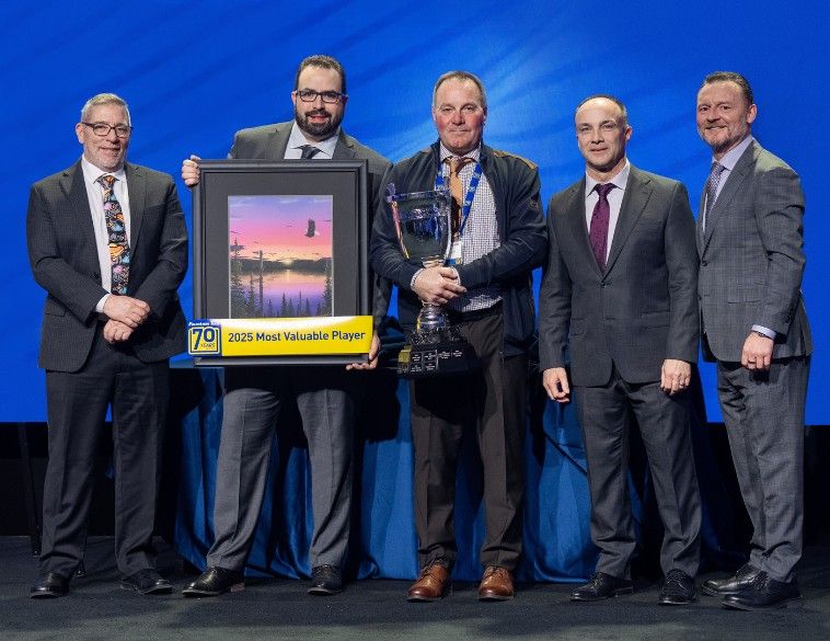 Brian Adcock, propriétaire de Fountain Tire d’Innisfail, se tient sur scène avec quatre autres hommes, tenant un grand trophée en argent. À ses côtés, un dirigeant tient un prix encadré de « Joueur le plus utile 2025 » arborant un logo « 70 ans. » Ce prix a été remis lors du congrès des propriétaires de Fountain Tire.