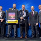 Brian Adcock, owner of Fountain Tire Innisfail, stands on stage with four other men, holding a large silver trophy. Beside him, an executive holds a framed "2025 Most Valuable Player" award featuring a "70 Years" logo, presented during the Fountain Tire Owners’ Convention.