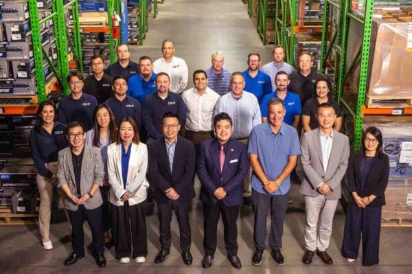 A group portrait of the GSP North America team standing in a large warehouse distribution center. The diverse group of professionals, wearing business casual and GSP-branded clothing, is positioned between tall green storage racks stocked with automotive parts. The setting highlights GSP’s commitment to providing local support and a deep understanding of regional needs.