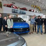 Une photo de groupe à l'intérieur de la concession Dubois Méthot Chevrolet à Victoriaville. Huit hommes posent fièrement de chaque côté d'un Chevrolet Equinox EV bleu éclatant. Au centre, Jean-François Dubois et le gagnant Dave Fortier se serrent la main devant le véhicule. En arrière-plan, sur une plateforme surélevée, on aperçoit des voitures de collection rouges.