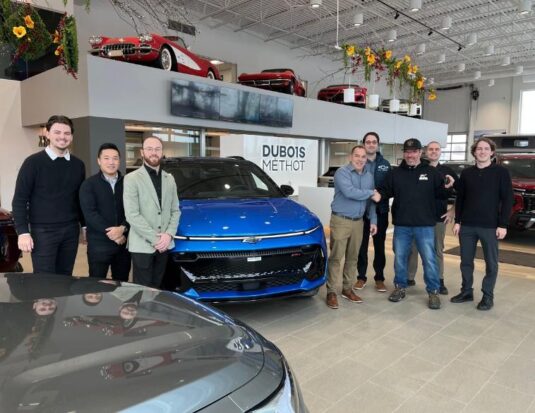 Une photo de groupe à l'intérieur de la concession Dubois Méthot Chevrolet à Victoriaville. Huit hommes posent fièrement de chaque côté d'un Chevrolet Equinox EV bleu éclatant. Au centre, Jean-François Dubois et le gagnant Dave Fortier se serrent la main devant le véhicule. En arrière-plan, sur une plateforme surélevée, on aperçoit des voitures de collection rouges.