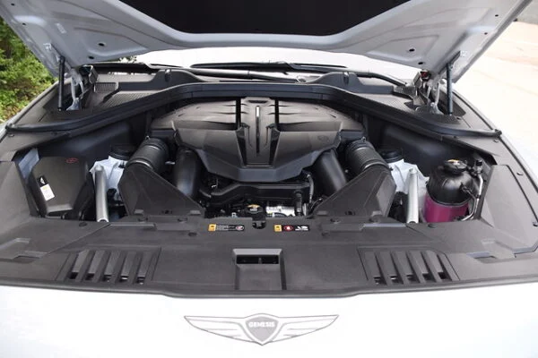 A wide shot of the open engine bay of a silver Genesis vehicle, featuring a large black protective engine cover and a complex, tightly packed layout of various automotive components.