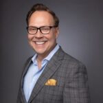 Portrait de Travis Parman, nouveau directeur des communications chez Goodyear, portant un complet à carreaux gris et des lunettes.