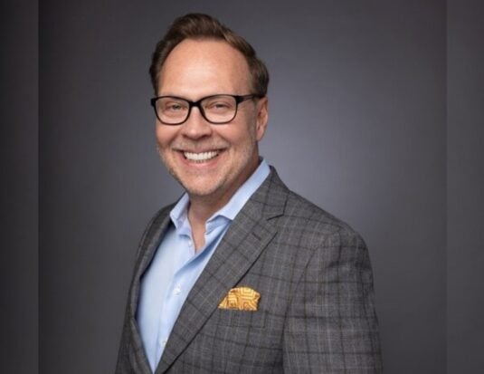 Portrait de Travis Parman, nouveau directeur des communications chez Goodyear, portant un complet à carreaux gris et des lunettes.