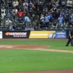 Une vue de l'avant-champ d'un stade de baseball, montrant une rangée de panneaux publicitaires sur la clôture. Les panneaux incluent '414 Day', une grande publicité 'Hankook Tire' avec 'driving emotion' et le logo 'Dynapro', et une publicité pour 'Carbliss'. Un arbitre marche sur la terre battue devant la clôture. Des spectateurs sont visibles dans les gradins derrière.