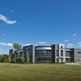 Exterior view of the Nexen Tire America Technical Center, a modern two-story corporate building featuring a dark grey masonry and glass facade with a distinctive curved glass corner. The building is situated on a well-manicured green lawn under a clear blue sky, with the Nexen Tire logo prominently displayed on the structure and a smaller sign near the road.