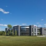 Vue extérieure du centre technique de Nexen Tire America, un bâtiment d'entreprise moderne de deux étages avec une façade en maçonnerie gris foncé et en verre, doté d'un angle vitré incurvé distinctif. Le bâtiment est situé sur une pelouse verdoyante sous un ciel bleu clair, avec le logo Nexen Tire bien en évidence sur la structure et un panneau plus petit près de la route.