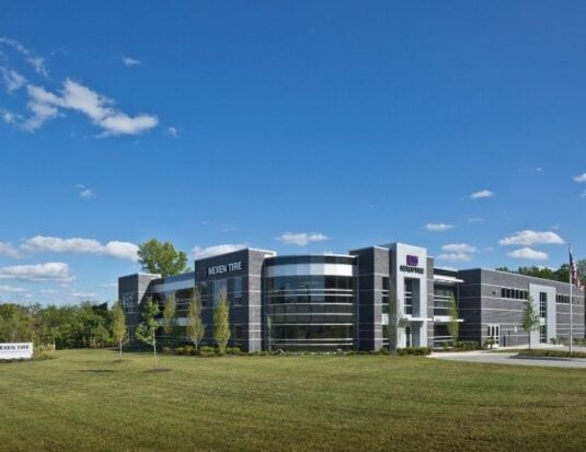 Vue extérieure du centre technique de Nexen Tire America, un bâtiment d'entreprise moderne de deux étages avec une façade en maçonnerie gris foncé et en verre, doté d'un angle vitré incurvé distinctif. Le bâtiment est situé sur une pelouse verdoyante sous un ciel bleu clair, avec le logo Nexen Tire bien en évidence sur la structure et un panneau plus petit près de la route.