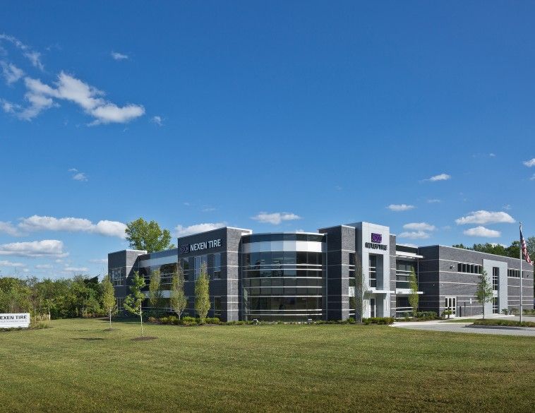 Vue extérieure du centre technique de Nexen Tire America, un bâtiment d'entreprise moderne de deux étages avec une façade en maçonnerie gris foncé et en verre, doté d'un angle vitré incurvé distinctif. Le bâtiment est situé sur une pelouse verdoyante sous un ciel bleu clair, avec le logo Nexen Tire bien en évidence sur la structure et un panneau plus petit près de la route.