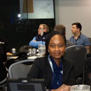 Portrait d'Inès Tchakima, directrice des communications et des relations publiques d'Auto Prévention, assise à une table lors du colloque 2026. Elle porte un veston noir sur une blouse bleue à nœud, avec un cordon d'identification autour du cou. Une tablette est posée devant elle et d'autres participants sont visibles en arrière-plan dans la salle de conférence.