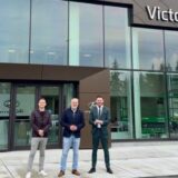 Three men, Jordan Kot, Vice President, John Kot, President, and Brandon Kot, Vice President, from left to right, pose for a photograph in front of the Victoria Hyundai dealership, according to the Kot Auto Group and CNW Group. They are standing on a concrete walkway in front of the modern, glass-fronted building.