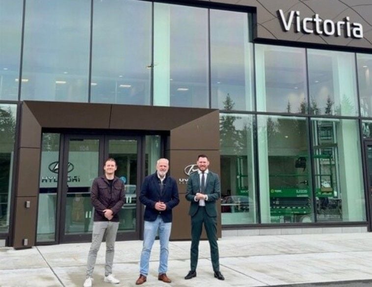 Three men, Jordan Kot, Vice President, John Kot, President, and Brandon Kot, Vice President, from left to right, pose for a photograph in front of the Victoria Hyundai dealership, according to the Kot Auto Group and CNW Group. They are standing on a concrete walkway in front of the modern, glass-fronted building.