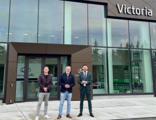Trois hommes, Jordan Kot, vice-président, John Kot, président, et Brandon Kot, vice-président, de gauche à droite, posent pour une photographie devant le concessionnaire Victoria Hyundai, selon le Kot Auto Group et le Groupe CNW. Ils se tiennent sur une passerelle en béton devant le bâtiment moderne à la façade vitrée.