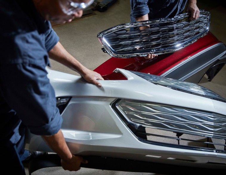 Two technicians in a workshop handle automotive body parts; one person holds a white front bumper assembly while another person in the background holds a chrome mesh grille.