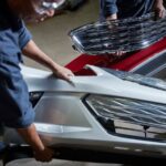 Deux techniciens en carrosserie manipulant des pièces de véhicule, dont un couvre-pare-chocs blanc au premier plan et une calandre chromée à l'arrière-plan.
