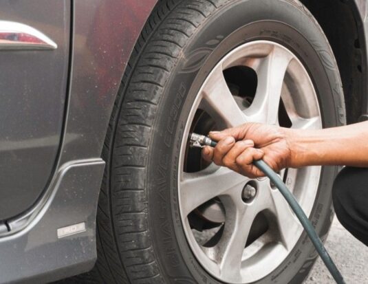 A close-up view of a person's hand attaching a compressed air hose to the valve stem of a car tire mounted on a silver alloy rim.