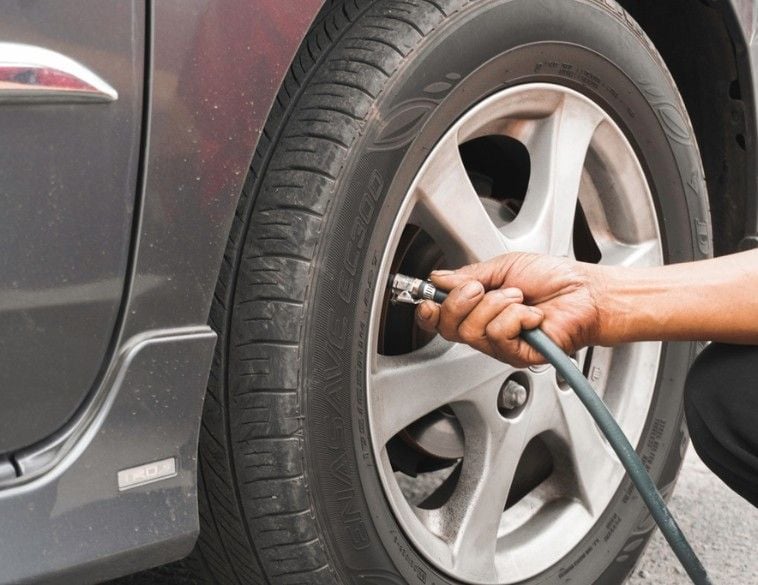 A close-up view of a person's hand attaching a compressed air hose to the valve stem of a car tire mounted on a silver alloy rim.