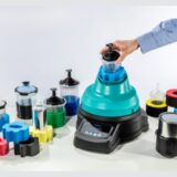 A person’s hand places a mixing cup into the turquoise and black ROTOGEN 2 car refinish paint mixer, which is surrounded by various spray gun cup systems and colorful foam adapters on a white surface.