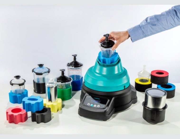 A person’s hand places a mixing cup into the turquoise and black ROTOGEN 2 car refinish paint mixer, which is surrounded by various spray gun cup systems and colorful foam adapters on a white surface.