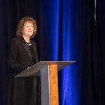 Sylvie Mallette, directrice générale d'Auto Prévention, prend la parole derrière un pupitre lors d'un colloque annuel de sensibilisation à la santé et à la sécurité au travail. Elle porte un collier de perles et un micro-casque. Le fond est un rideau bleu foncé plissé.