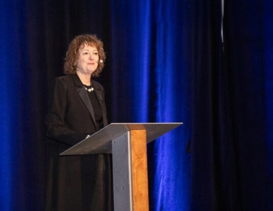 Sylvie Mallette, directrice générale d'Auto Prévention, prend la parole derrière un pupitre lors d'un colloque annuel de sensibilisation à la santé et à la sécurité au travail. Elle porte un collier de perles et un micro-casque. Le fond est un rideau bleu foncé plissé.