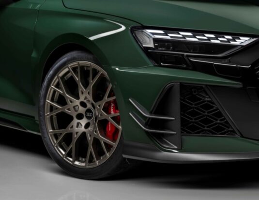 Close-up of a dark green Audi RS 3 competition limited featuring a bronze multi-spoke alloy wheel, red Audi ceramic brake calipers, and Pirelli P Zero Trofeo R tires. The image highlights the carbon fiber aero canards and modern LED headlight design.