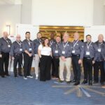 Un groupe de treize professionnels de l’industrie, la plupart portant des chemises grises uniformisées avec des cordons d’identification, posant pour une photo d'équipe devant une entrée de salle de bal au Hilton Mississauga/Meadowvale.