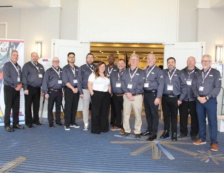 Un groupe de treize professionnels de l’industrie, la plupart portant des chemises grises uniformisées avec des cordons d’identification, posant pour une photo d'équipe devant une entrée de salle de bal au Hilton Mississauga/Meadowvale.