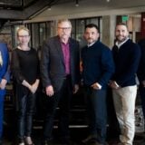 A group of six professionally dressed people from PBS Systems and Dealer Image Pro standing side-by-side in a modern automotive showroom, posing together to commemorate their partnership.